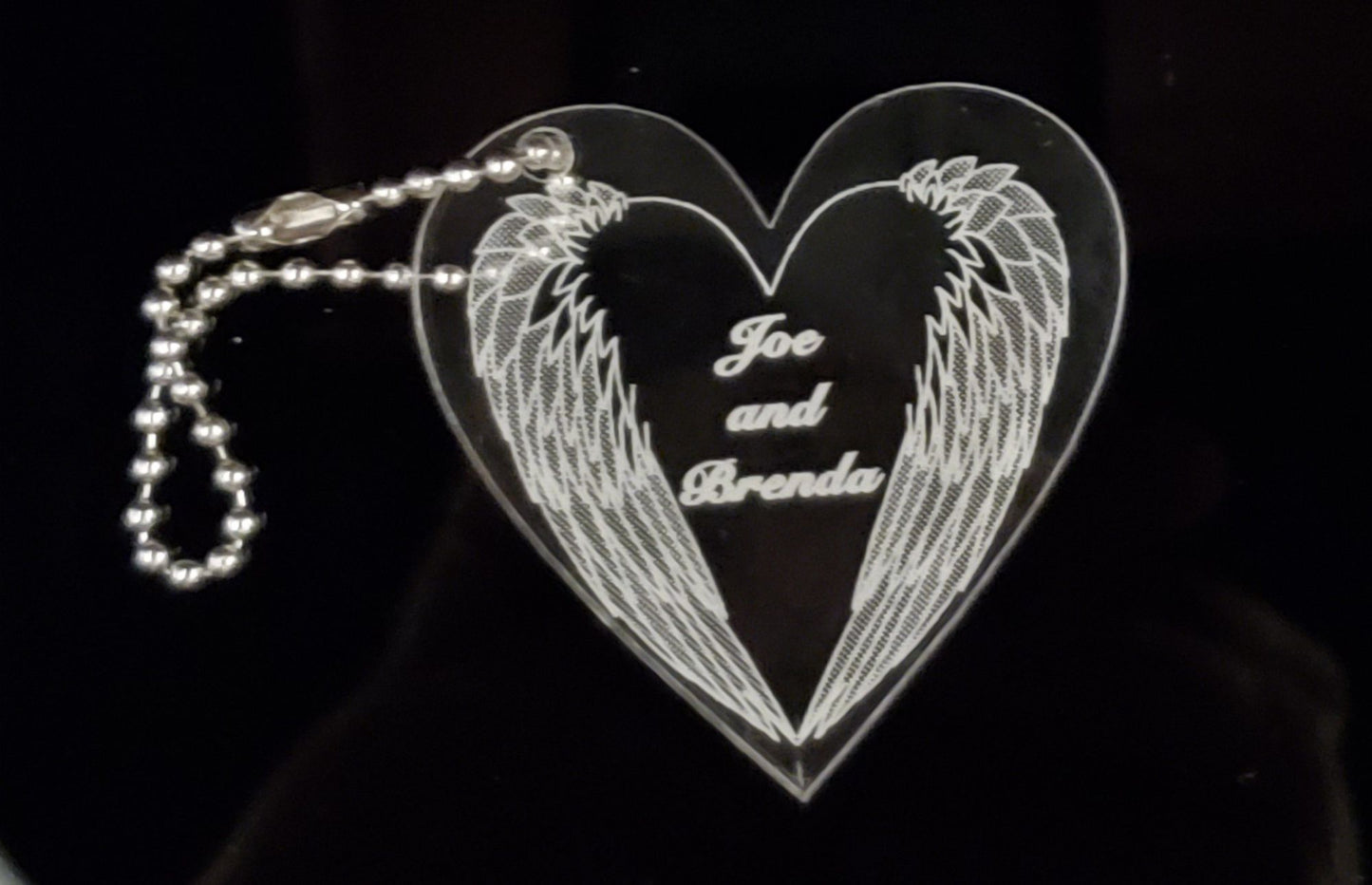 heart shaped acrylic keychain designed with a downward angel wings and engraved with names, with a small metal chain attached