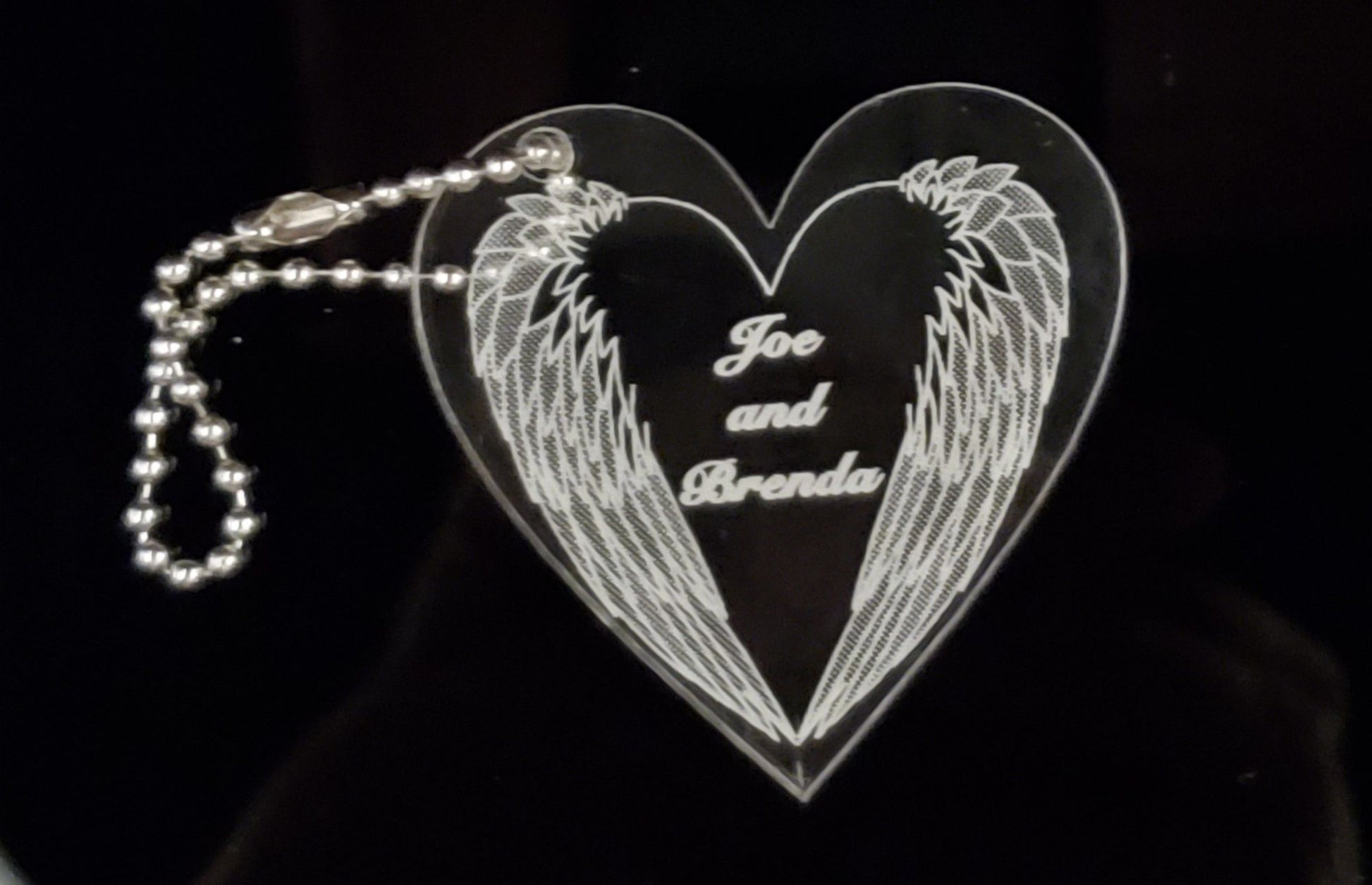 heart shaped acrylic keychain designed with a downward angel wings and engraved with names, with a small metal chain attached