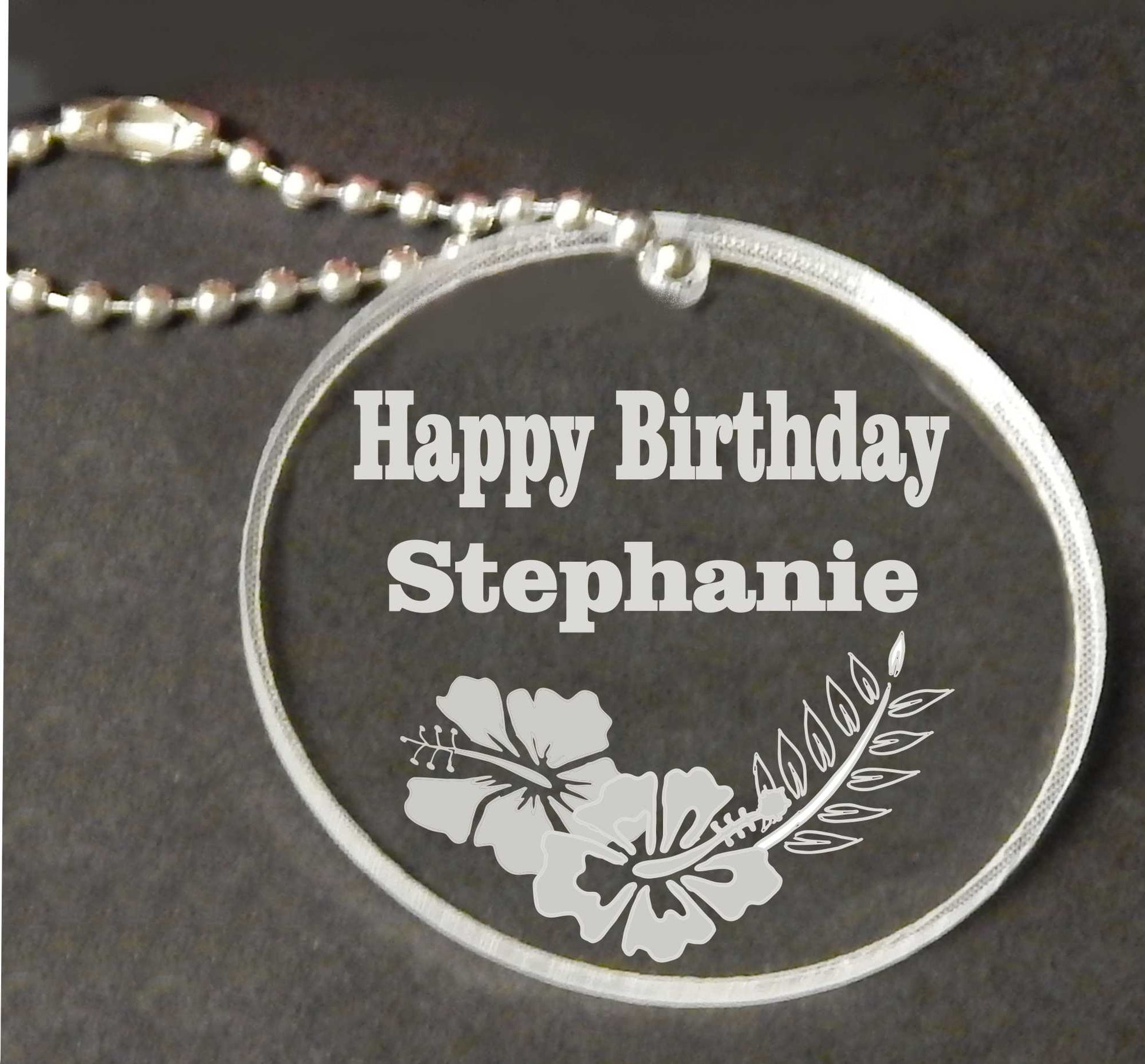 clear acrylic round keychain shown with Happy Birthday, name and designed with hibiscus flowers at bottom along with a small chain attached