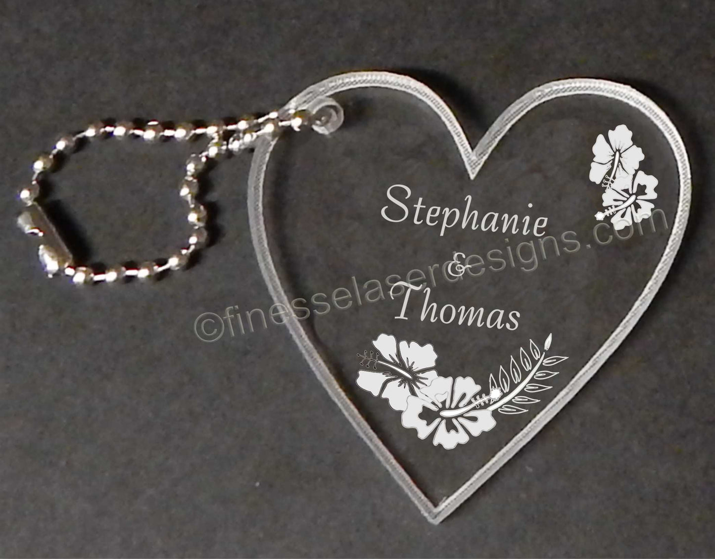 an acrylic heart shaped key chian with an hibisuc flower design and names engraved with a small metal chain attached