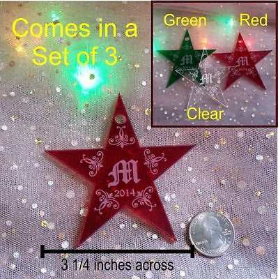 acrylic red star shaped ornament with a monogrammed letter M in the center of the ornament next to a quarter to show size dimension and then a corner square showing the ornament in clear acrylic, red acrylic and green acrylic