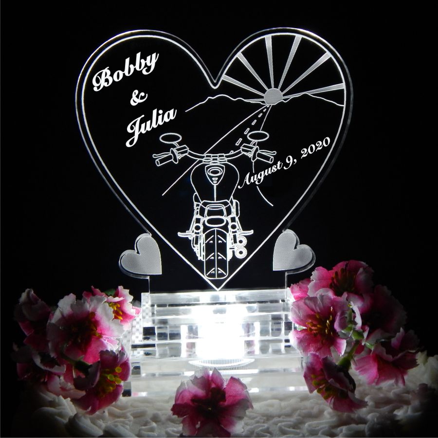 Acrylic heart shaped cake topper with backside view of motorcycle driving on road towards a sunset with names and date